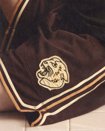 A close-up of Bobcat Shorts, brown 1970s-style basketball shorts featuring a roaring bobcat patch and gold striped trim.