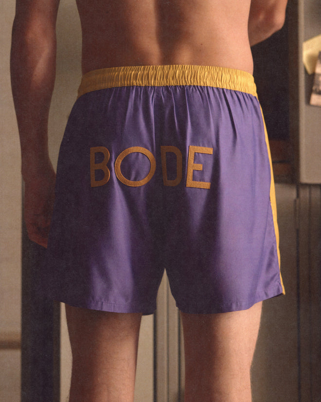 Someone stands facing away, wearing the Satin Field Shorts - Purple, featuring purple and yellow panels, a contrasting waistband, and "BODE" displayed on the back.