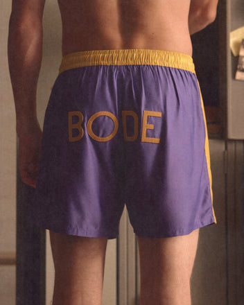 Someone stands facing away, wearing the Satin Field Shorts - Purple, featuring purple and yellow panels, a contrasting waistband, and 