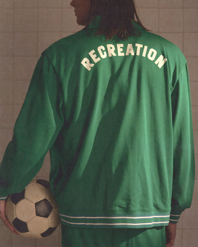 Person in a green Sophomore Jacket with "RECREATION" on the back, holding a soccer ball and facing away—features heavyweight fabric and vintage jersey styling for an authentic sporty look.