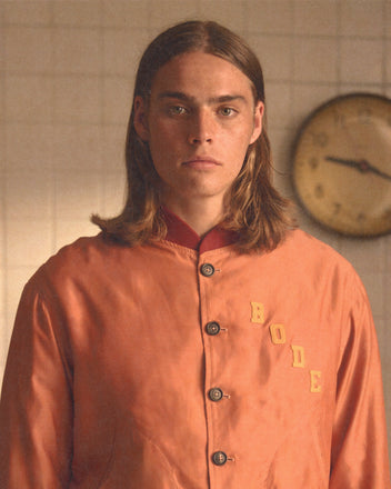 A person with long hair wears a peach Rally Jacket, inspired by 1960s varsity styles, standing in front of a wall clock.