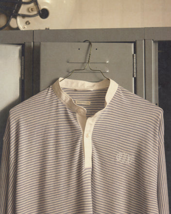 The Newton Stripe Polo - Blue, a striped long-sleeve polo with a band collar, hangs on a wire hanger in front of a gray locker.