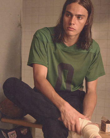 A young person with long brown hair wears a green Scrimmage Jersey, sitting indoors and gazing thoughtfully to the side.