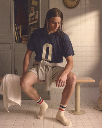 A young man wearing the Scrimmage Jersey - Navy and athletic gear sits on a locker room bench, looking down, with a towel draped beside him.