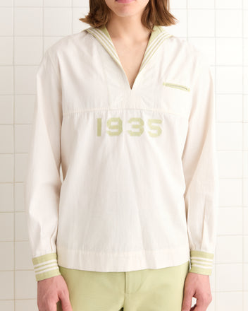 A person in a white 1935 Cricket Shirt featuring a distinctive sailor collar stands against a tiled wall.