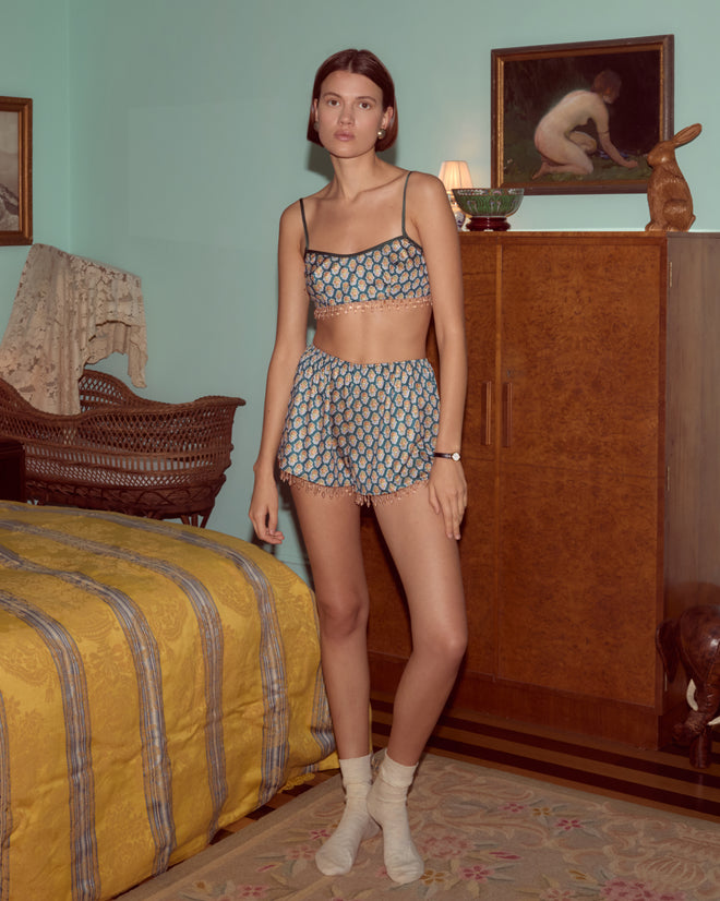 A woman wearing Beaded Bouquet Shorts and socks stands in a vintage-style bedroom with yellow bedding and wooden furniture.
