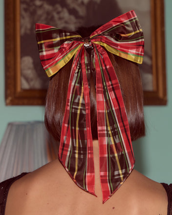 Someone with short brown hair wears the Carol Bell Bow—a large, one-of-a-kind vintage plaid ribbon featuring a brass bell charm—at the back of their head.