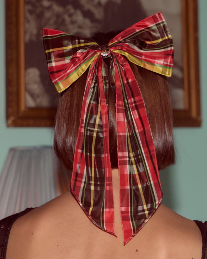 Someone with short brown hair wears the Carol Bell Bow—a large, one-of-a-kind vintage plaid ribbon featuring a brass bell charm—at the back of their head.