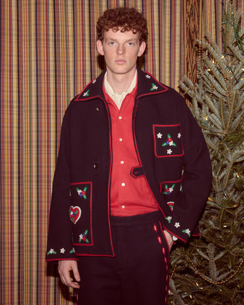 A young man wearing the Carroll Front Tab Jacket, decorated with heart and flower appliqués, stands by a Christmas tree and striped curtains.