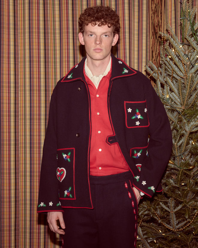 A young man wearing the Carroll Front Tab Jacket, decorated with heart and flower appliqués, stands by a Christmas tree and striped curtains.