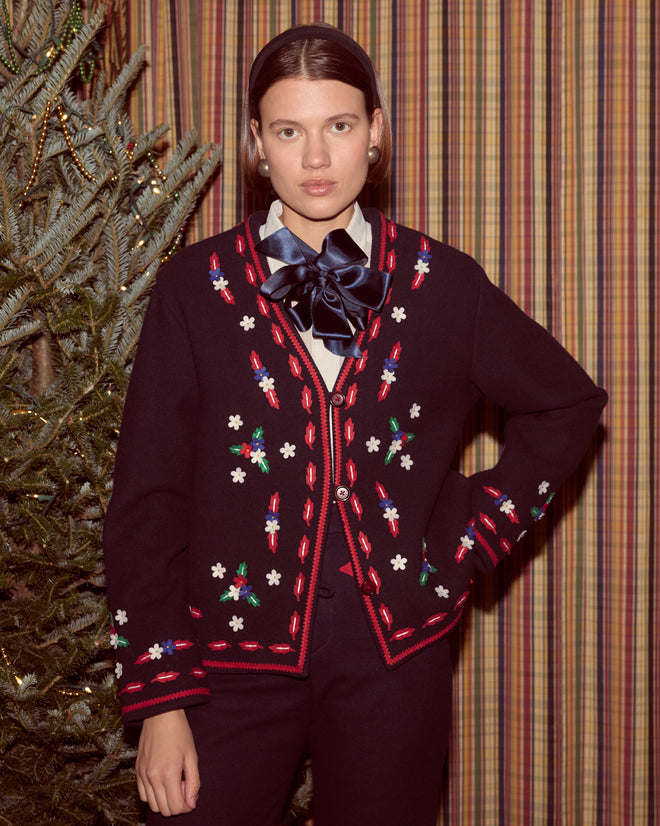 A woman in the Carroll Jacket with floral appliqué stands by a Christmas tree and striped curtains, her black cardigan elegantly embroidered.