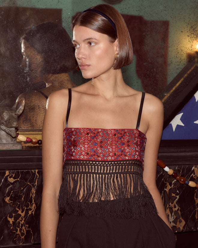 A woman with a bob haircut wears the Cloister Garden Bra - S, a unique red embroidered crop top with black fringe, standing indoors by a mirror.