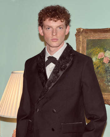 A young man in a Beaded Connaught Tuxedo Jacket stands indoors beside a lamp and a framed floral painting.