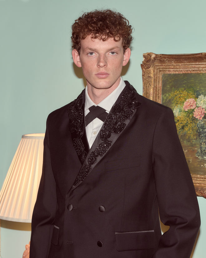 A young man in a Beaded Connaught Tuxedo Jacket stands indoors beside a lamp and a framed floral painting.