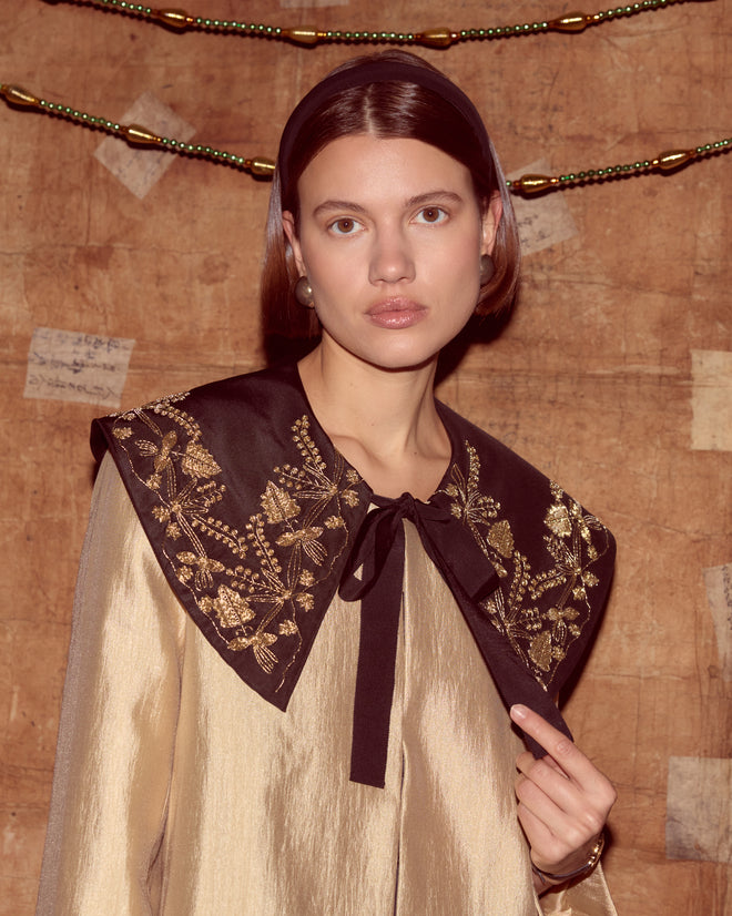 A woman wears the Gilt Jacket—a gold piece with a black embroidered collar inspired by Ottoman Empire styles—standing before a textured brown wall adorned with beads.