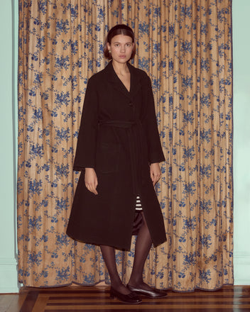 A woman stands indoors on a wooden floor in front of floral curtains, wearing the Hemlock Coat—a long black wool coat with a belted waist.
