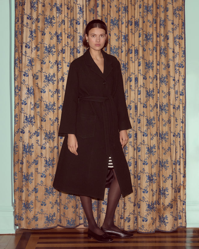 A woman stands indoors on a wooden floor in front of floral curtains, wearing the Hemlock Coat—a long black wool coat with a belted waist.