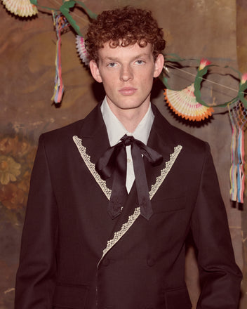 A young man with curly hair wears the Lacework Double-Breasted Tuxedo Jacket and a bow tie, standing before festive decor.