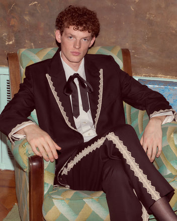 Man with curly red hair in a black suit and Lacework Side-Buckle Trousers sits on a patterned armchair, looking at the camera.