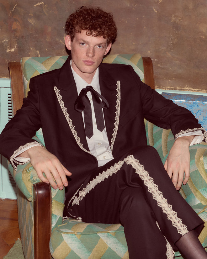 Man with curly red hair in a black suit and Lacework Side-Buckle Trousers sits on a patterned armchair, looking at the camera.