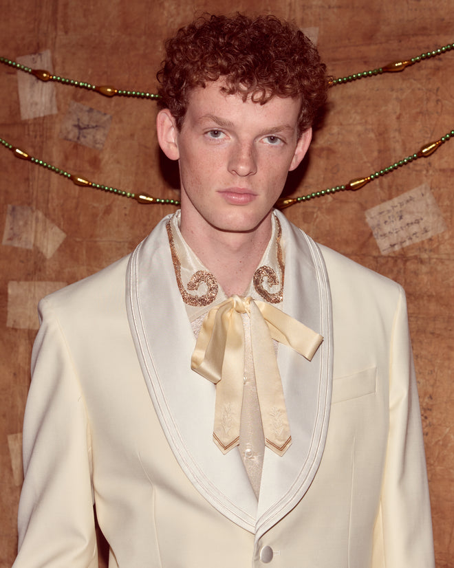 A young man with curly red hair wears the Meringue Shawl Collar Tuxedo Jacket with a shiny bowtie as he stands before a rustic backdrop.