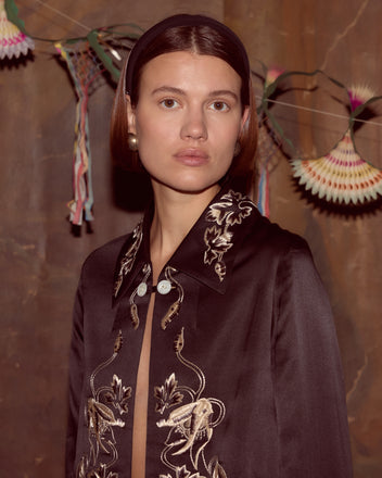 A woman wearing the Phantasm Fringe Jacket - Black stands in front of paper fans and ribbons on a brown wall.