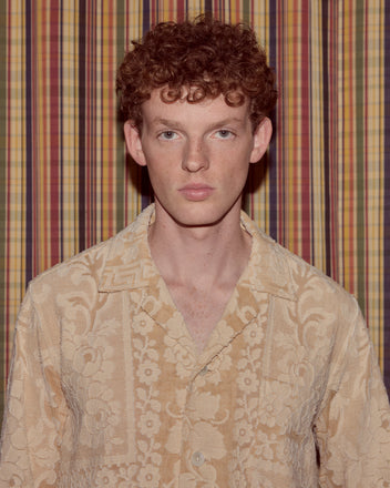 A young person with curly red hair, wearing the Sandstone Key Overshirt in size S, stands before a striped, multicolored curtain.