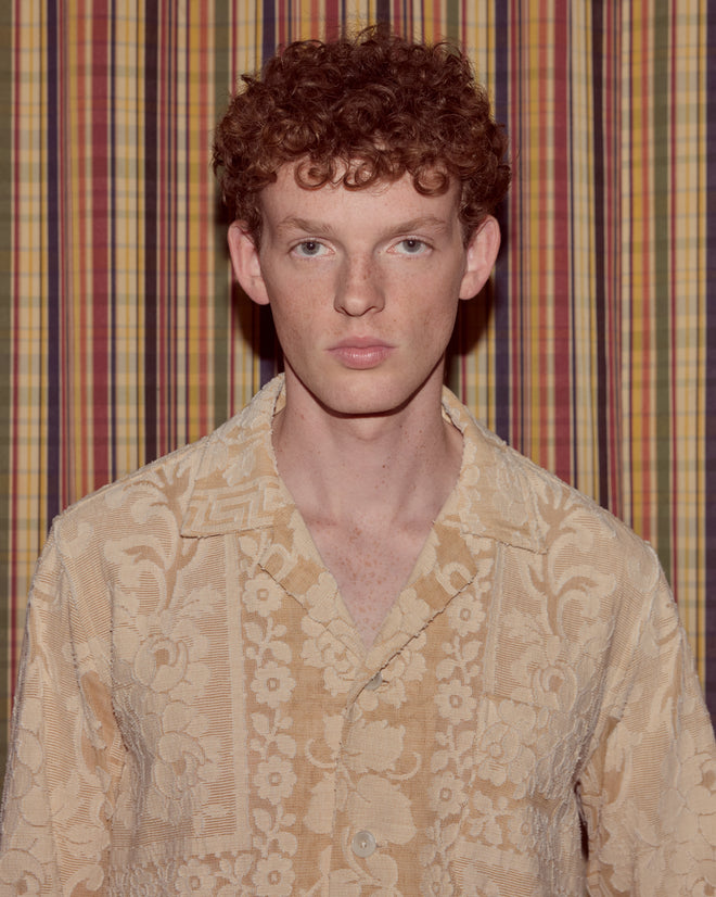 A young person with curly red hair, wearing the Sandstone Key Overshirt in size S, stands before a striped, multicolored curtain.