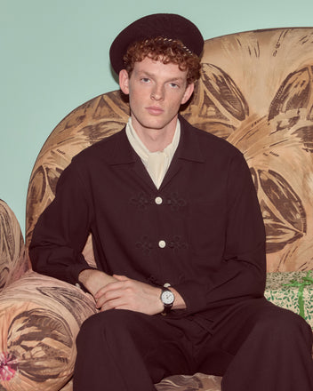A man with curly hair sits on a patterned armchair, wearing the Astor Trillium Shirt in black and a watch, looking at the camera.