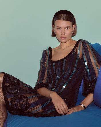 A woman in a Sequin Podium Dress sits on a blue couch with a light teal background.