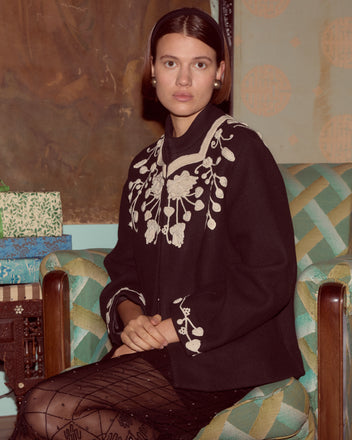 A woman wearing the Snowdrop Appliqué Jacket sits on a patterned chair, looking calmly at the camera.