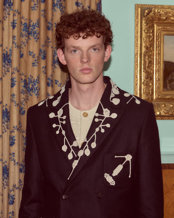 A man with curly hair wears the Snowdrop Appliqué Suit Jacket in navy, featuring white embroidered designs, standing indoors by a curtain.