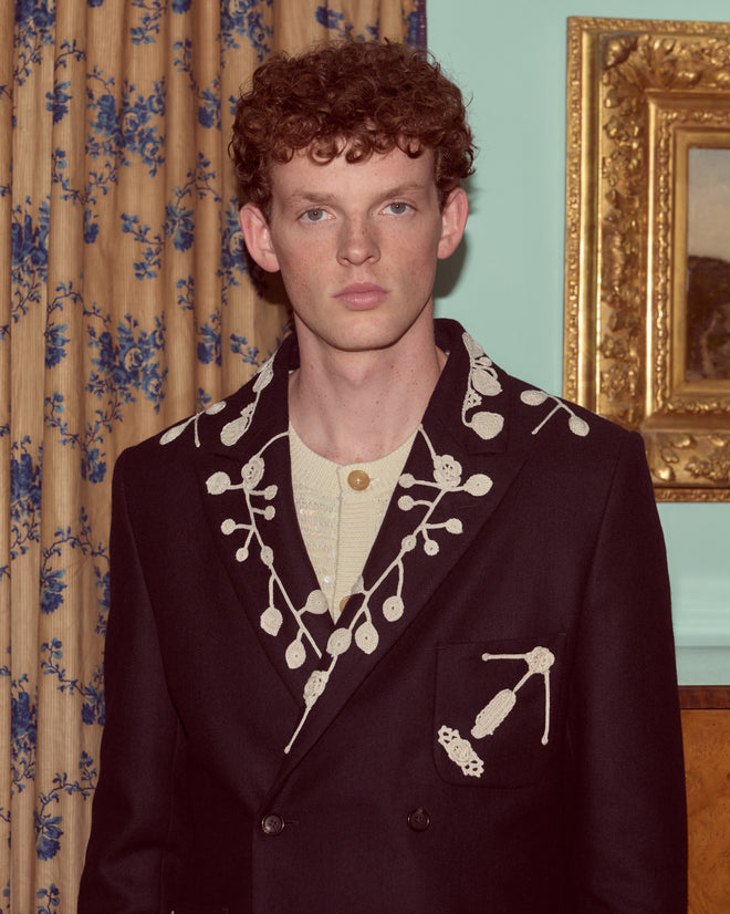 A man with curly hair wears the Snowdrop Appliqué Suit Jacket in navy, featuring white embroidered designs, standing indoors by a curtain.