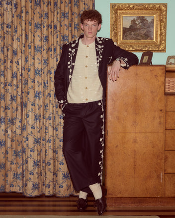 A man in embroidered black suit and Snowdrop Appliqué Trousers leans on a wooden cabinet in a vintage room with floral curtains.