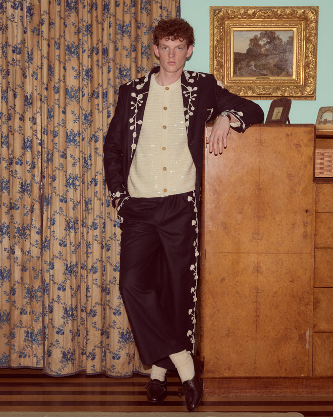 A man in embroidered black suit and Snowdrop Appliqué Trousers leans on a wooden cabinet in a vintage room with floral curtains.