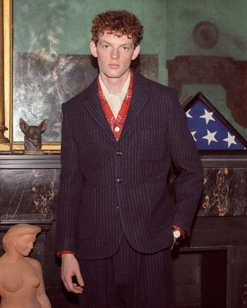 A man in a Waldorf Pinstripe Suit Jacket stands by a mantle, with a folded US flag and statues displayed behind him.