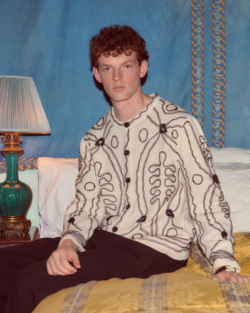 A man with curly hair sits on a bed beside a lamp, wearing the patterned Winter Garden Cardigan and black pants.