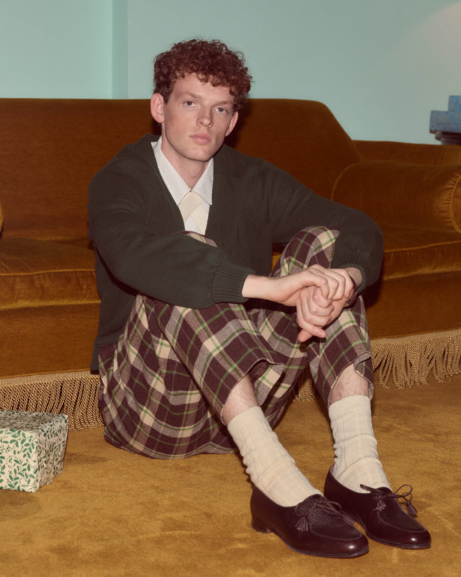 A young man with curly hair sits on a brown carpet wearing Woodcrest Trousers and a cozy sweater, in front of a vintage sofa.