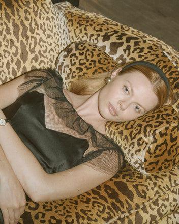 A woman in the Prima Dress, a black silk gown with an illusion neckline, reclines on a leopard-print couch and gazes neutrally at the camera.