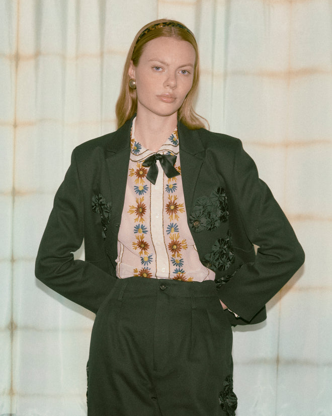 A woman in the Ribbon Garland Jacket - Black, paired with a floral blouse and headband, stands before light-patterned curtains.