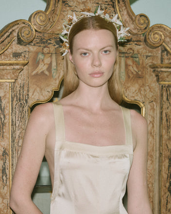 A woman with straight blonde hair, wearing the Loulou Jumpsuit in cream and a floral headpiece, stands before an ornate wood panel.