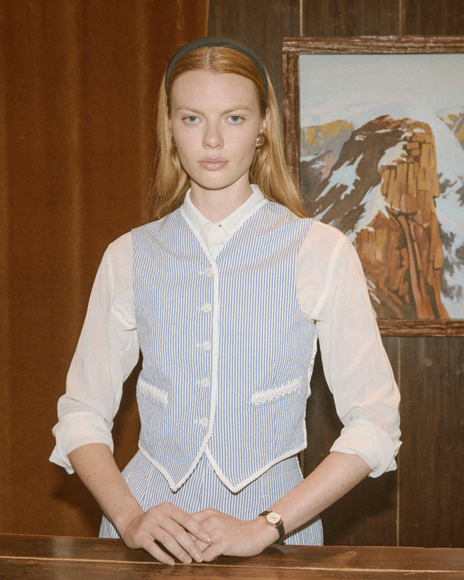 A woman in the Club Stripe Waistcoat - Blue/White stands at a desk with a mountain painting on the wall behind her.