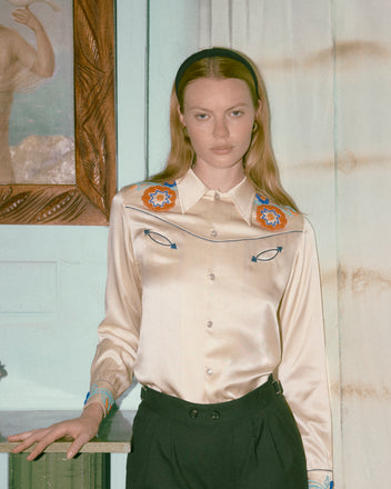 Woman in a satin blouse with eye designs and Tropical Wool Double Button Shorts, standing indoors before a curtain and framed painting.
