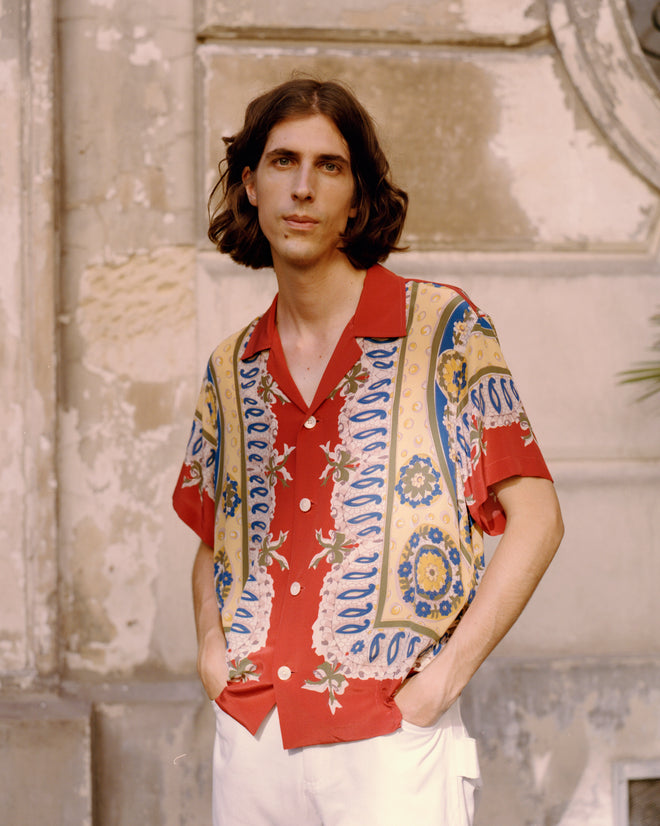 A person with shoulder-length hair wears the vibrant Bricolage Short Sleeve Shirt and stands with hands in pockets against a weathered stone wall.