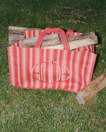 A Parasol Stripe Tote filled with firewood rests on green grass, with scattered logs nearby.