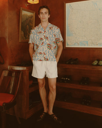 A person wearing the Swimmers Short Sleeve Shirt and white shorts stands indoors by shoe shelves and a U.S. map, highlighting this classic midcentury style.