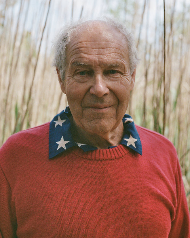 An older man wears the Juniper Sweater in pink/red—a washable merino wool knit—over a blue shirt with white stars, standing outdoors among tall grasses.