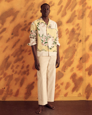 A person stands barefoot against an orange patterned backdrop, wearing light-colored pants and the Jasmine Sketch Shirt - L/XL, a unique yellow printed top with a floral pattern.