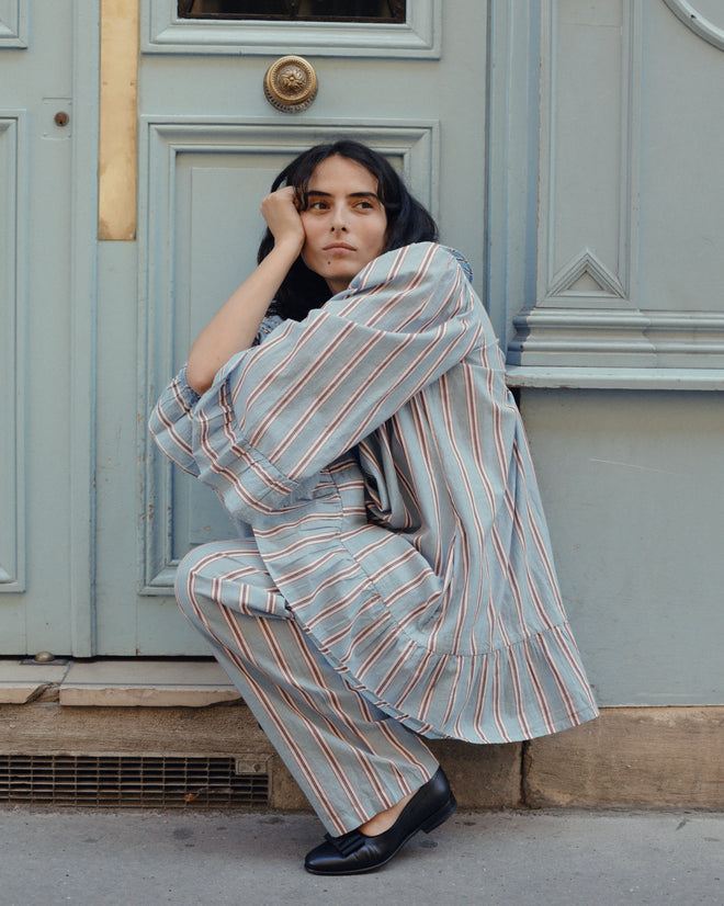 A woman in the Regatta Stripe Dress crouches by a blue door, resting her head on her hand with a thoughtful expression. The dress’s classic stripes and vintage silhouette recall the charm of 1930s shirting styles.