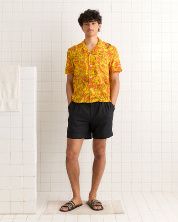 A man wearing the Abstract Foliage Short Sleeve Shirt, black shorts, and sandals stands in a white tiled room.